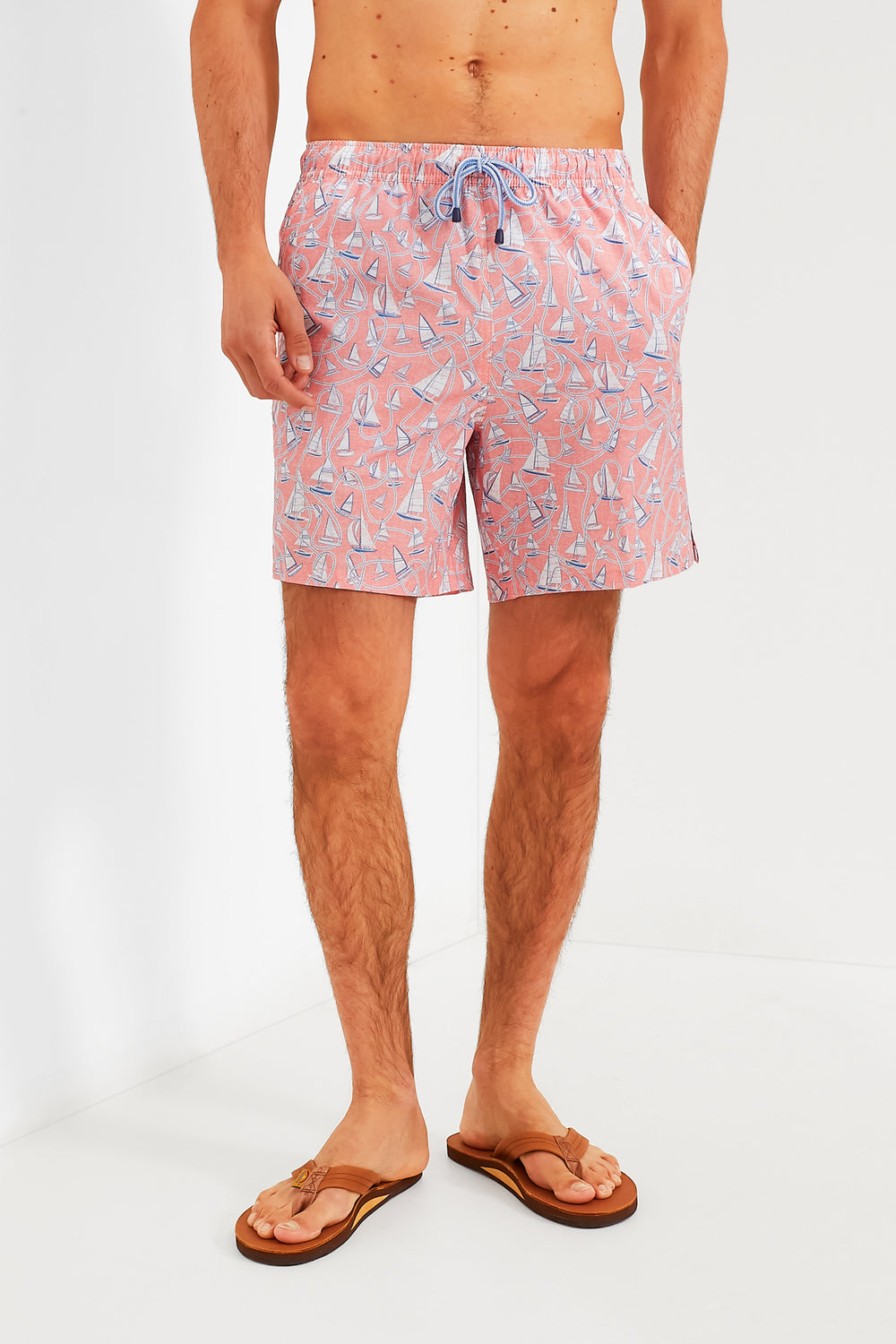 A man stands confidently in Peter Millar pink, quick-drying swim trunks with a sailboat pattern and beige flip-flops, featuring comfortable four-way stretch for easy movement.