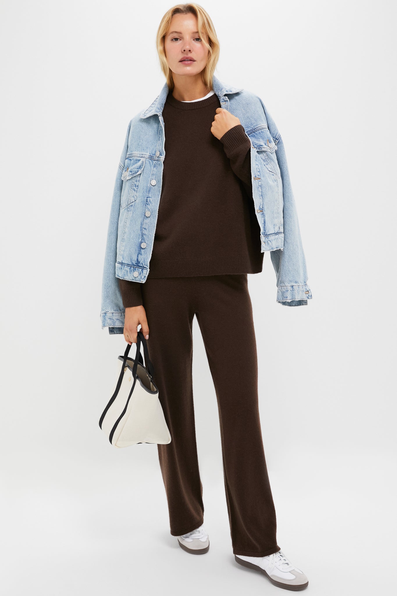 A woman wears a light denim jacket over a brown sweater with relaxed-fit cashmere pants, white sneakers, styled with a white handbag.