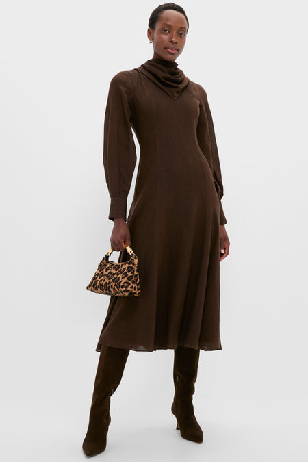 A woman in an Emilia Wickstead long-sleeve brown wool midi dress, knee-high brown boots, and holding a small leopard print handbag stands against a plain light background.