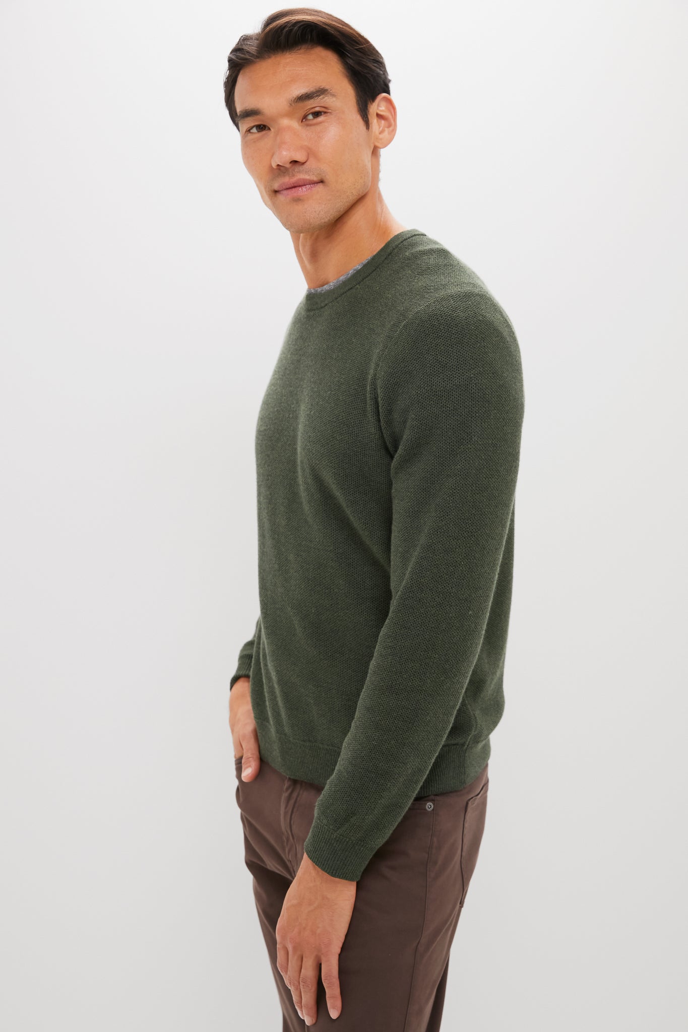 A man in a super-soft Marine Layer dark green sweater and brown pants stands against a plain white background, looking slightly toward the camera.