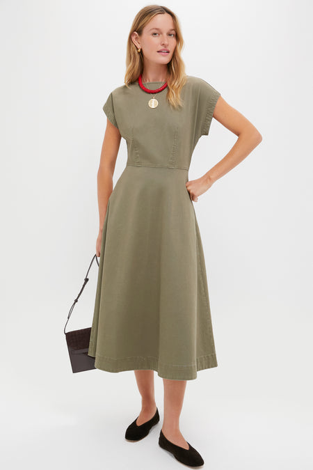 A woman in a Tuckernuck olive green short-sleeve corset-style midi dress with a fitted waist and A-line silhouette, black flats, a red necklace, and holding a black handbag stands against a plain backdrop.