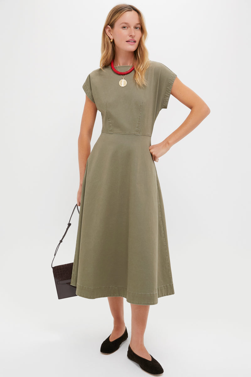 A woman in a Tuckernuck olive green short-sleeve corset-style midi dress with a fitted waist and A-line silhouette, black flats, a red necklace, and holding a black handbag stands against a plain backdrop.