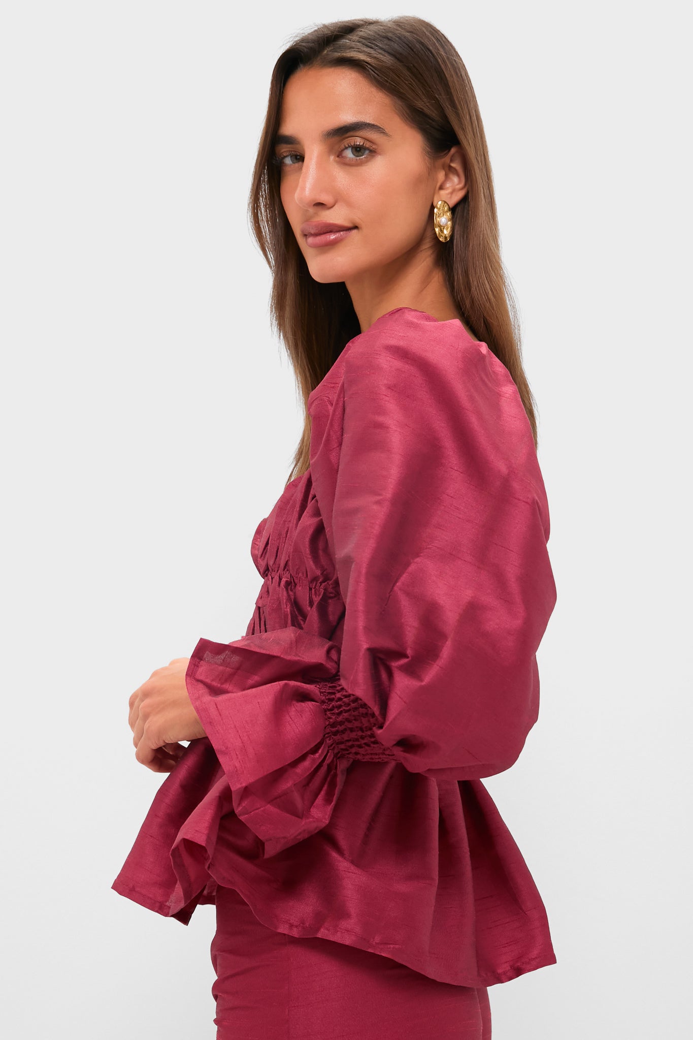 A woman wears the glamorous Louise Pink Mauve Elisabeth Top, a long-sleeved silk dupioni blouse with puffed sleeves, paired with gold earrings, posing against a plain light background.