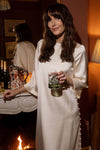 Woman in Winter White Side Button Jamie Dress, holding a drink by a marble mantel, exuding classic, effortless elegance with fun, high-end style; ideal for any occasion.