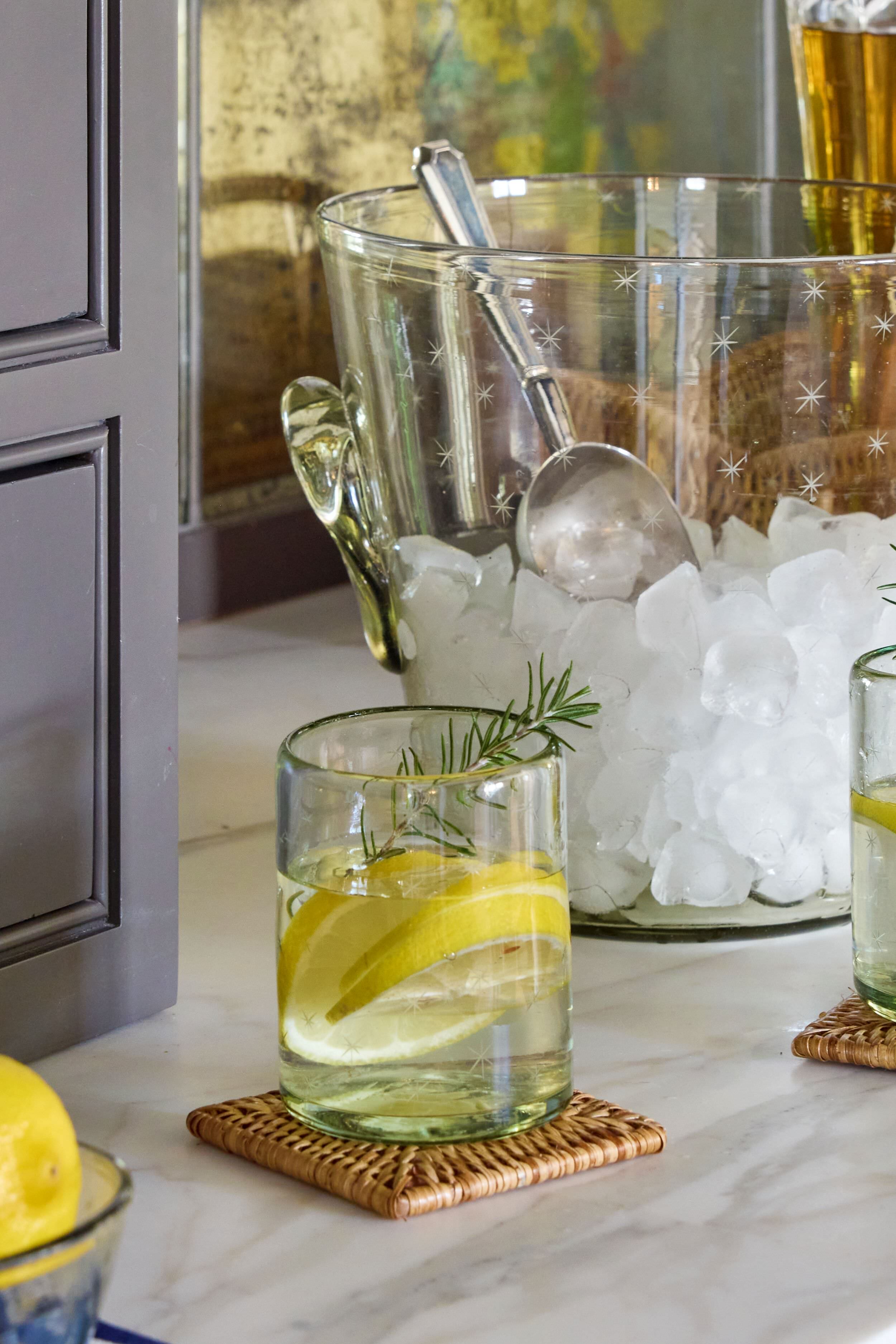 A glass of water with lemon slices and a rosemary sprig, served in handcrafted recycled glass by Half Past Seven, rests on a woven coaster beside a bowl of ice and a metal scoop.
