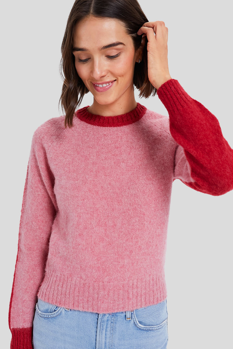 A woman smiles and touches her hair while wearing an Antik Batik color block knit pink sweater with red cuffs and collar, paired with light blue jeans.