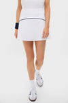 Dressed in Tnuck Sport’s white pleated-back tennis skirt with built-in compression shorts, a white top, athletic shoes, socks, and a navy wristband, the person stands confidently.