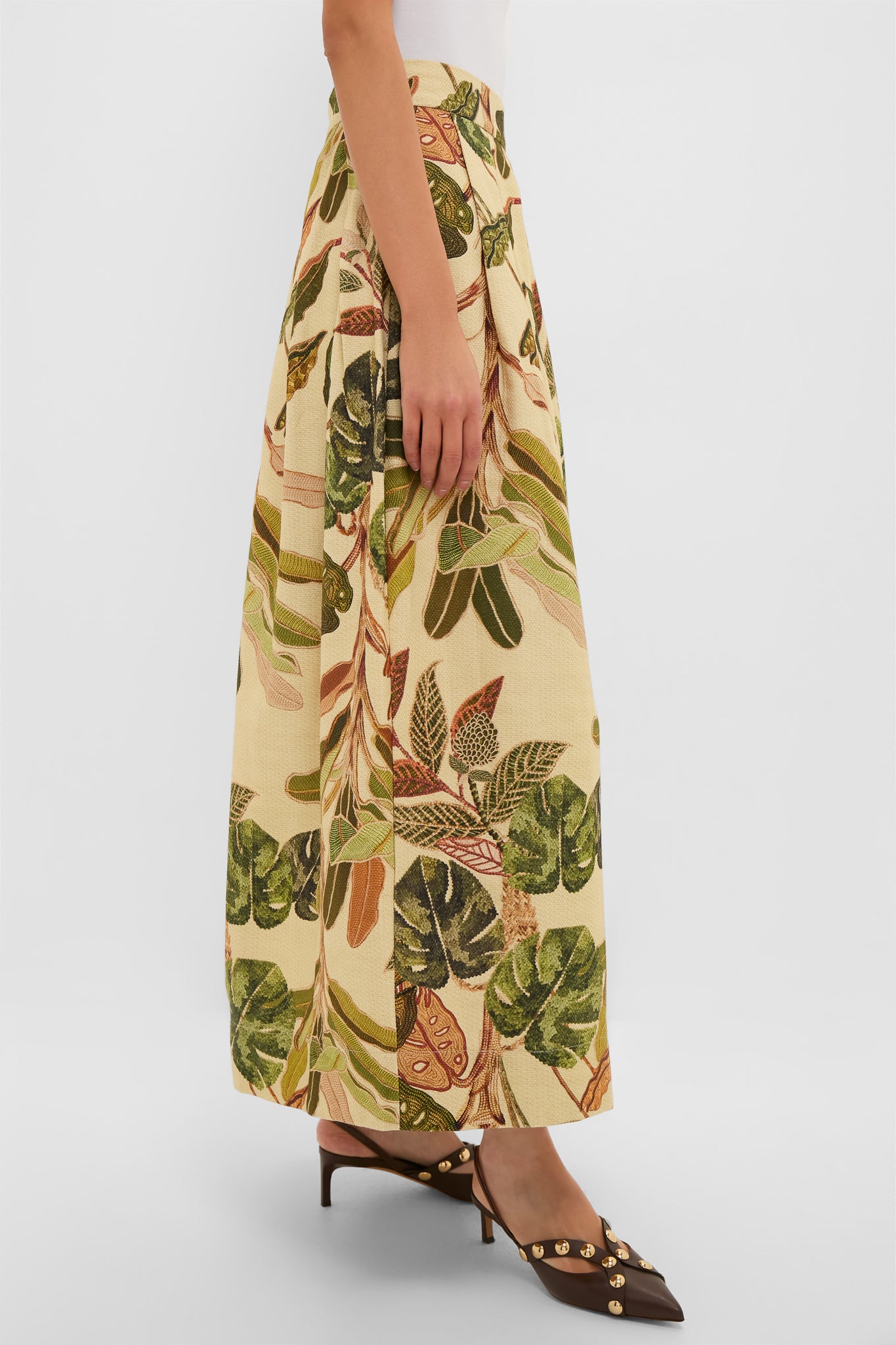 A person wearing a Dress To high-waisted, ankle-length skirt with tropical leaf print, a white top, and brown heeled shoes featuring gold embellishments.