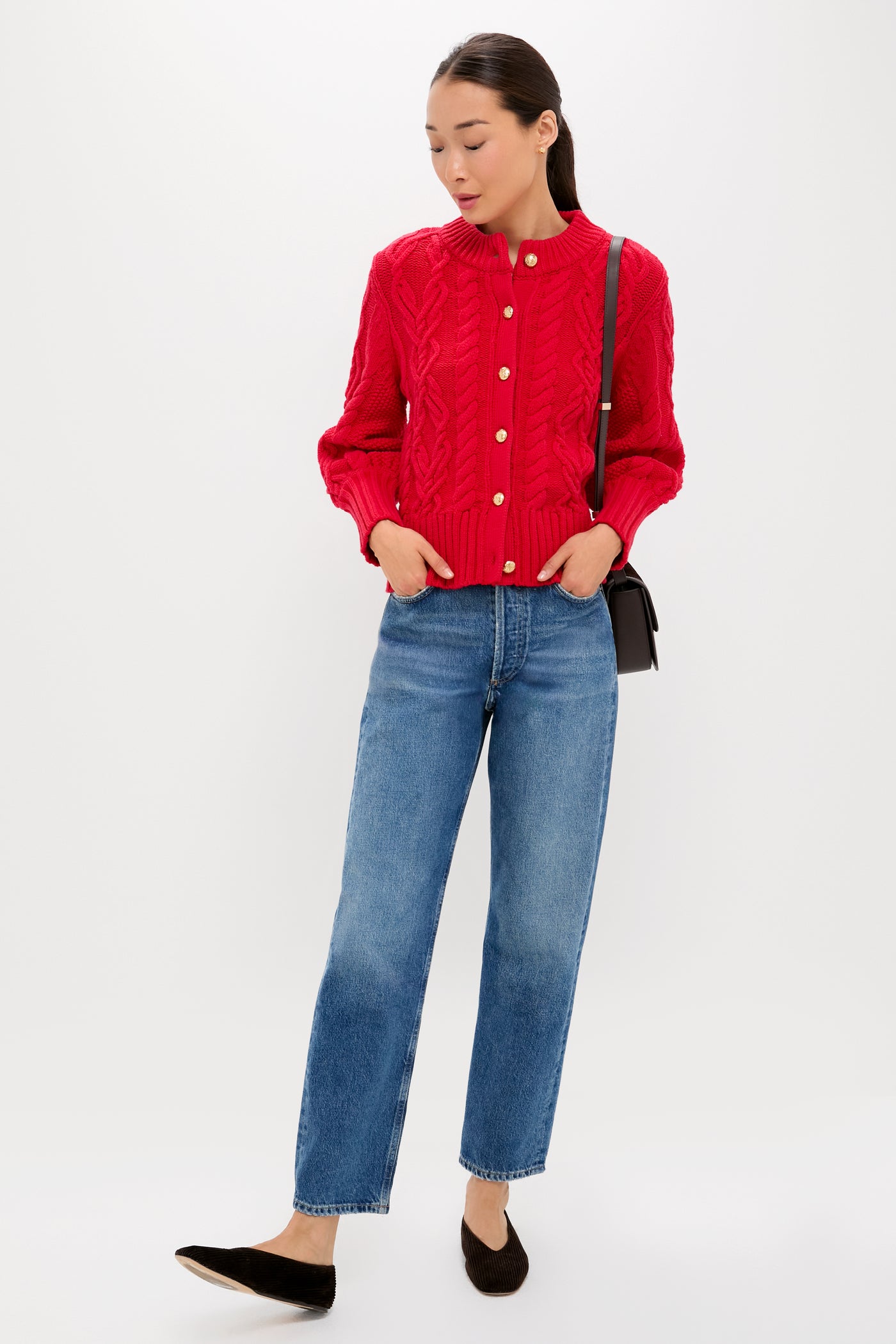 A woman wears a bright red chunky Tuckernuck cardigan, blue jeans, black flats, and a black crossbody bag, standing against a plain white background.