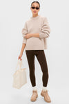 A person stands against a plain background wearing a beige sweater, brown leggings, socks with Stuart Weitzman suede boots, sunglasses, and holding a cream tote bag.