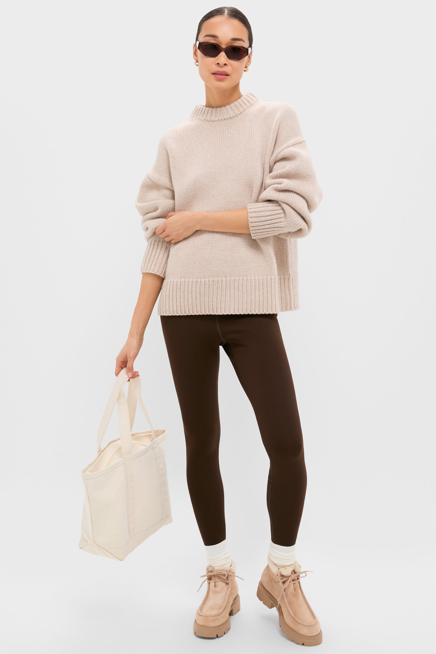 A person stands against a plain background wearing a beige sweater, brown leggings, socks with Stuart Weitzman suede boots, sunglasses, and holding a cream tote bag.