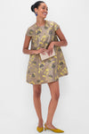 A woman in a shiny, vintage-inspired STAUD floral brocade mini dress holds a small clutch, wearing yellow pointed shoes and blue earrings, standing against a plain white background.