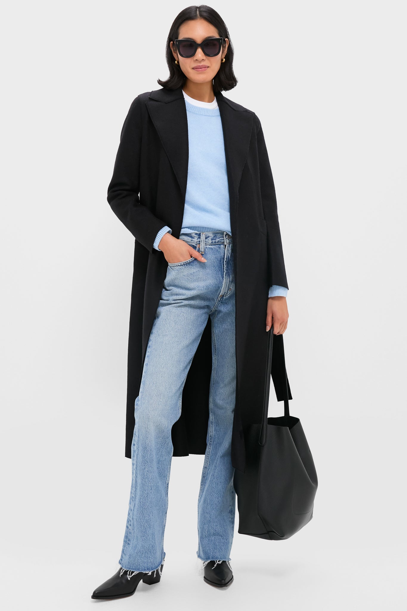 A person in a long black Dolce Vita coat, light blue sweater, blue jeans, black shoes, sunglasses, and carrying a large black tote bag stands against a plain background.