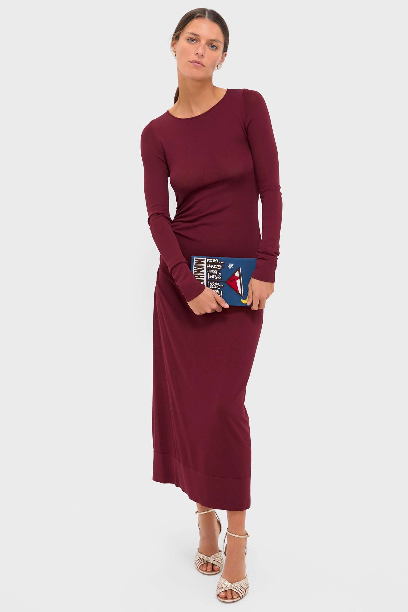 Model holding the Indigo Latim Manhattan Cocktail Book Clutch, a classic, hand-embroidered accessory, paired with an elegant burgundy dress and woven-strap heeled sandals. Effortless and high-end style.