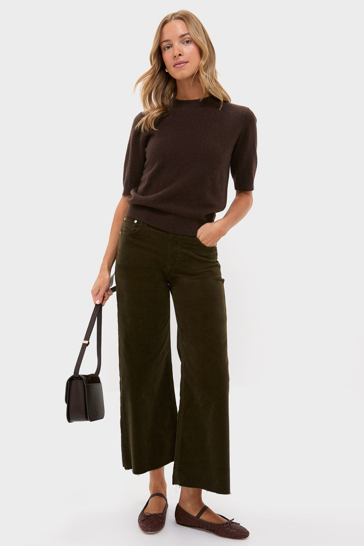 A woman stands against a white background wearing a dark brown short-sleeve sweater, Citizens of Humanity high-rise wide leg corduroy pants, brown flats, and holding a black shoulder bag.