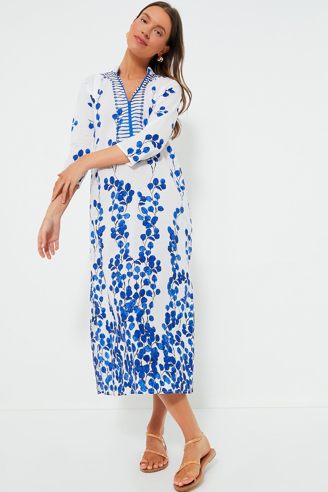 A woman wears a long white Ro's Garden Clorinda Kurta dress with blue floral patterns and gold sandals, made from 100% cotton—an ideal luxury travel-wear piece.