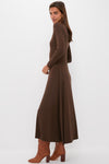 A woman stands sideways in a Tuckernuck Brown Sara Maxi knit dress with long sleeves and an ankle-length fit & flare silhouette, paired with brown heeled boots, against a plain white background.