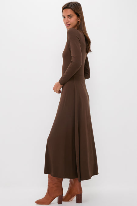 A woman stands sideways in a Tuckernuck Brown Sara Maxi knit dress with long sleeves and an ankle-length fit & flare silhouette, paired with brown heeled boots, against a plain white background.