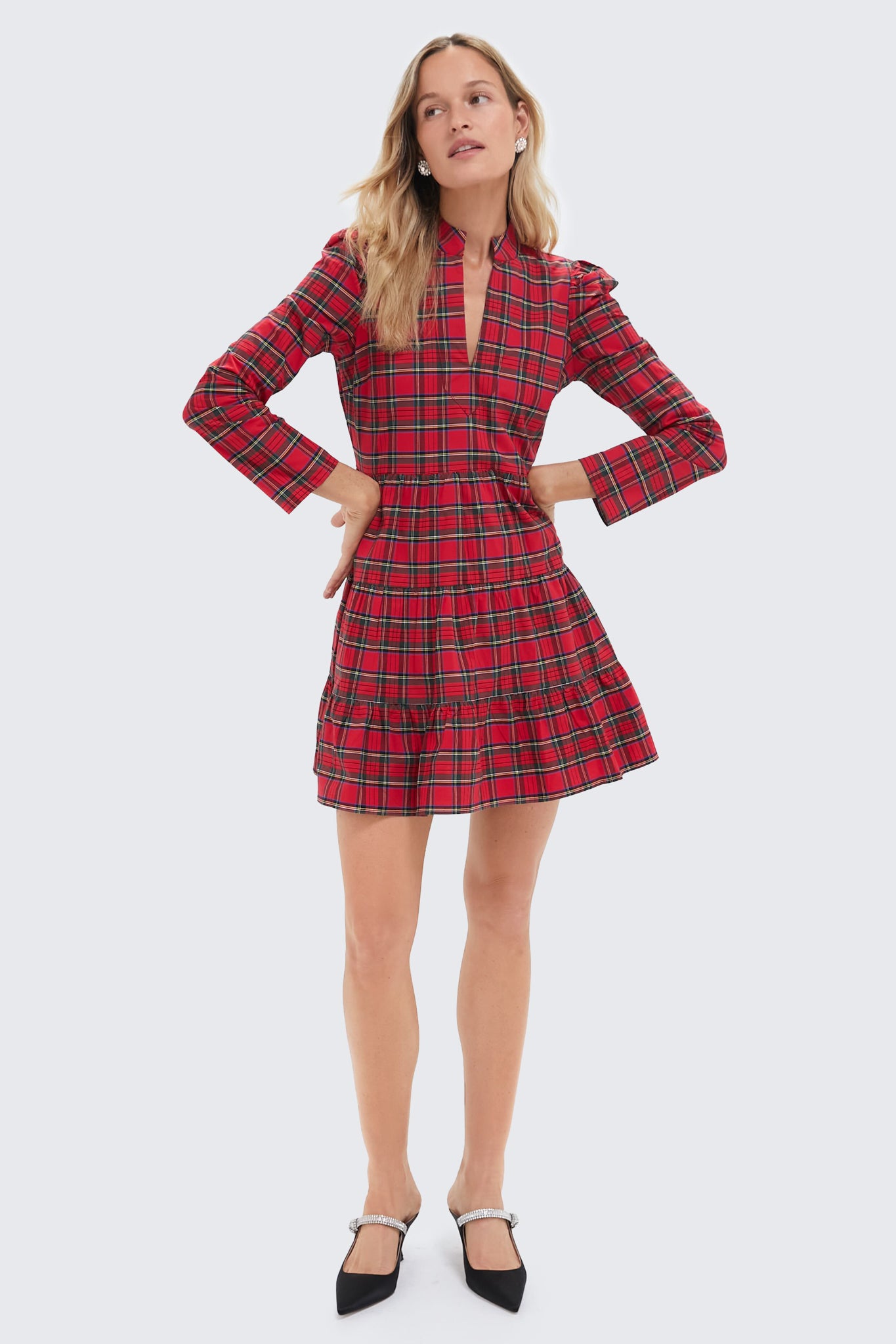 A woman in a Sail to Sable red plaid V-neck dress with a ruffled fit and flare tunic skirt and black pointed-toe heels stands against a plain light background.