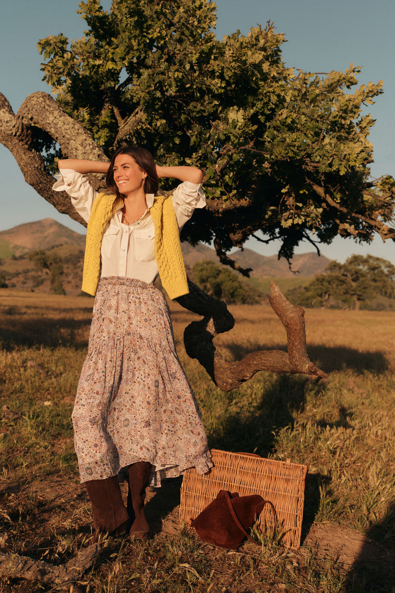 Woman standing outside in a long sleeved white button down shirt with the sleeves rolled up, a yellow cable knit sweater around her shoulders, a floral patterned maxi skirt, and chocolate brown boots. 