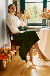 A woman in a Tuckernuck white button-down shirt with lantern sleeves and a dark green skirt sits by a window, holding a flower amid vases of flowers on the floor and table.