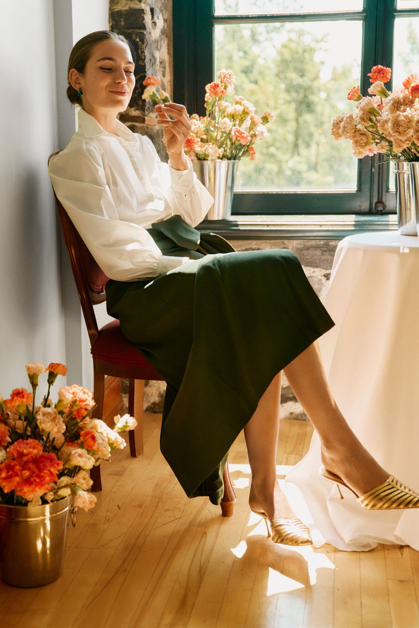 A woman in a Tuckernuck white button-down shirt with lantern sleeves and a dark green skirt sits by a window, holding a flower amid vases of flowers on the floor and table.