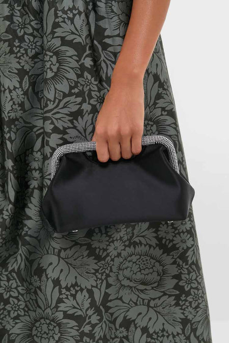 A person in a dark floral dress holds an Olga Berg evening clutch—a sleek black bag with a structured metallic frame—ideal for special occasions.