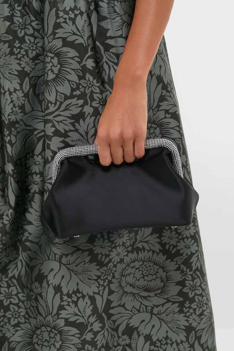 A person in a dark floral dress holds an Olga Berg evening clutch—a sleek black bag with a structured metallic frame—ideal for special occasions.
