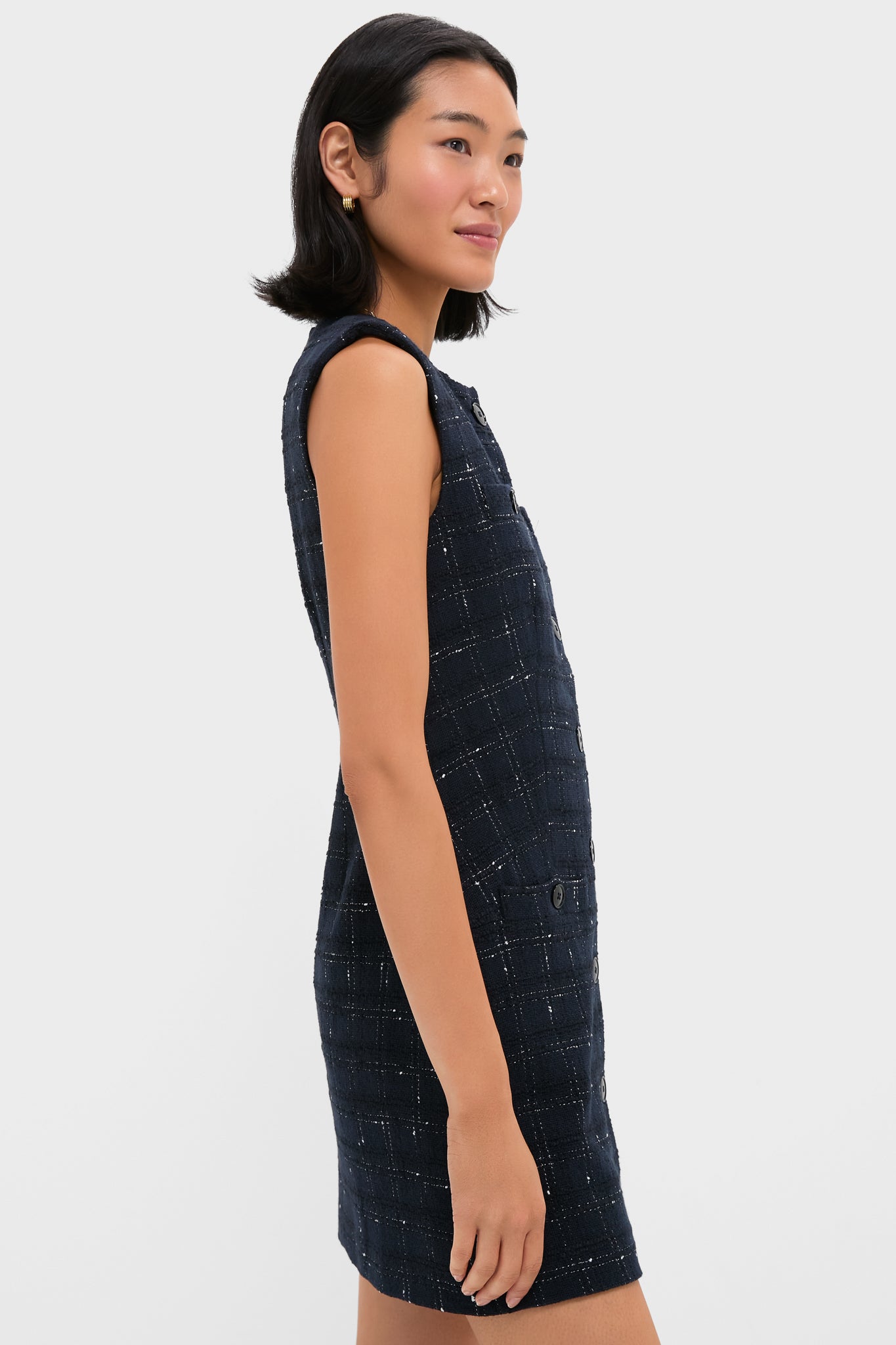 A woman stands in profile against a white background wearing a Veronica Beard sleeveless dark blue plaid mini dress with a structured silhouette and subtle check pattern.