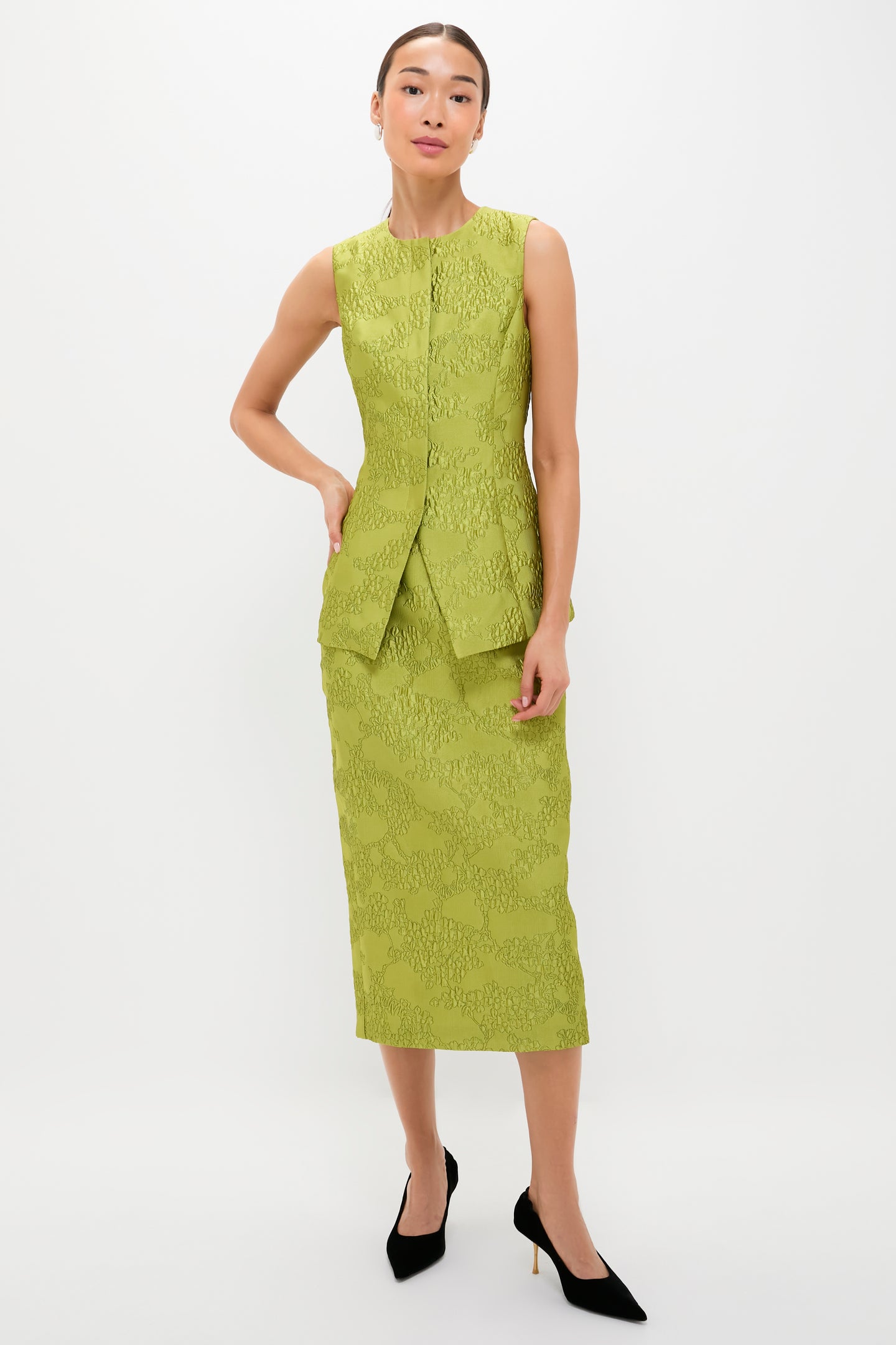 A person wears a sleeveless, textured light green jacquard midi skirt dress with a front slit from Tuckernuck and black pointed-toe heels, standing against a plain white background.