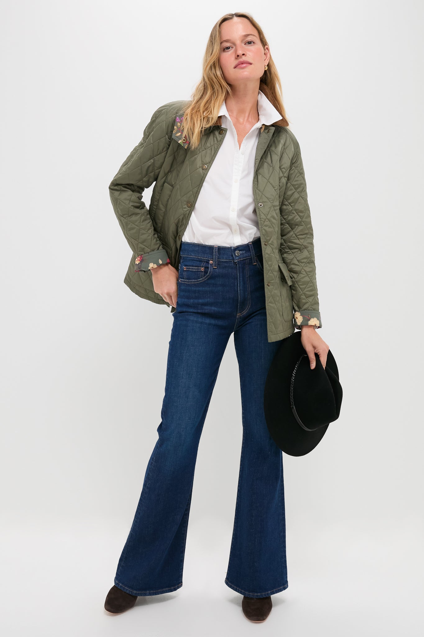 Model in olive quilted jacket, white shirt, and Zoe Gigi High Rise Skinny Flare jeans, holding a hat, embodies a classic, effortless, high-end style.