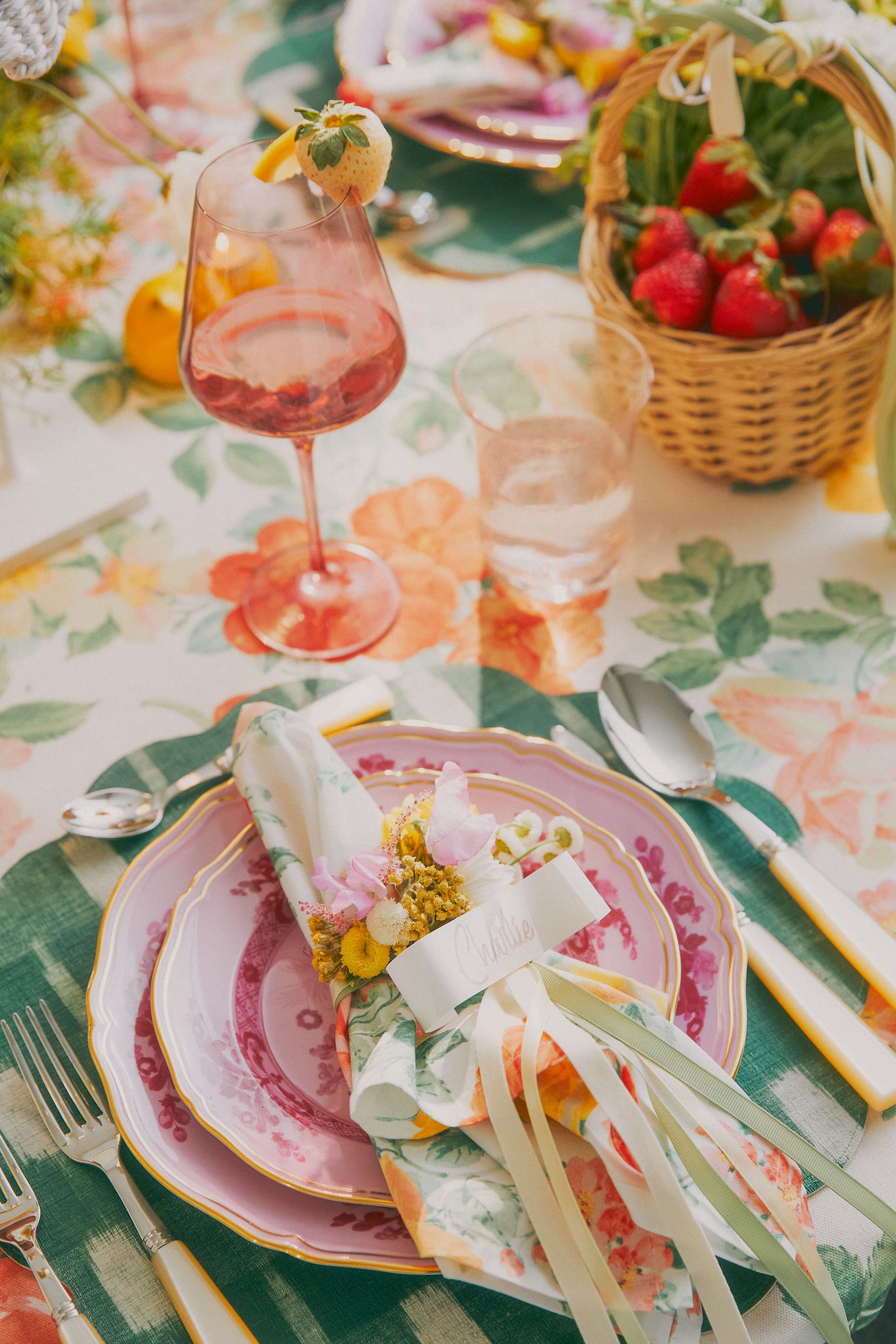 A floral table setting featuring layered porcelain plates, cutlery, wine and water glasses, a basket of strawberries, and a flower-adorned napkin with a name card evokes the elegant style of Ginori 1735.