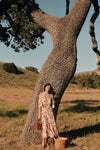 Woman standing in front of a tree in a a floral maxi dress with three quarter inch sleeves. She's holding a woven tan suede bag. 
