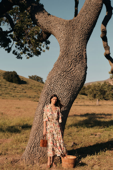 Woman standing in front of a tree in a a floral maxi dress with three quarter inch sleeves. She's holding a woven tan suede bag. 