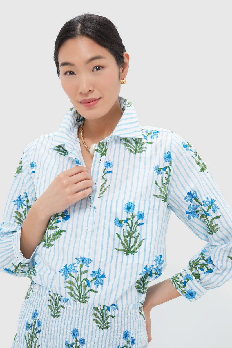 A woman wears a Pink City Prints blockprint shirt with blue floral designs, paired with gold earrings and a necklace. The handmade shirt is crafted in India from organic cotton. She stands touching her collar.