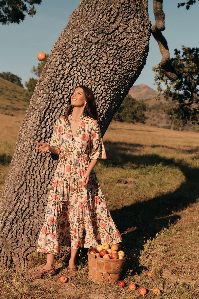 Woman standing in front of a tree throwing an apple in the air in a floral maxi dress. 