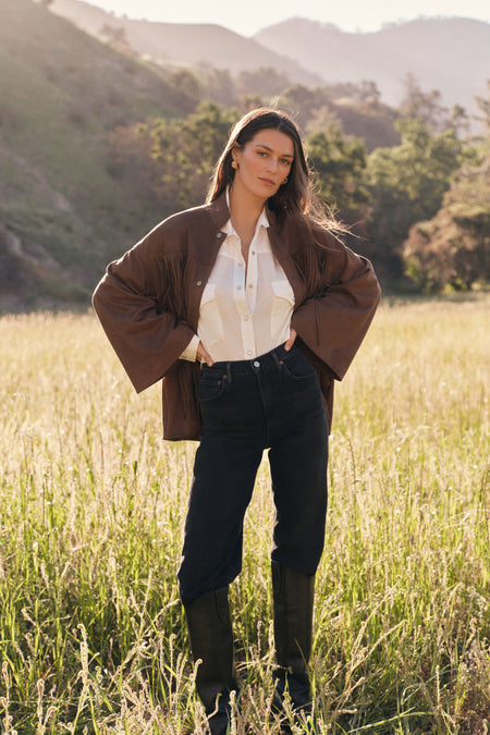 Woman in a brown fringe jacket, white button down shirt, dark wash jeans, and black leather boots. 