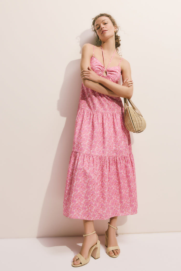 Woman in a tiered pink floral dress showcases the classic Natural Straw Malia Heels, featuring block heels and adjustable ankle straps, exuding an effortless, high-end summer style.