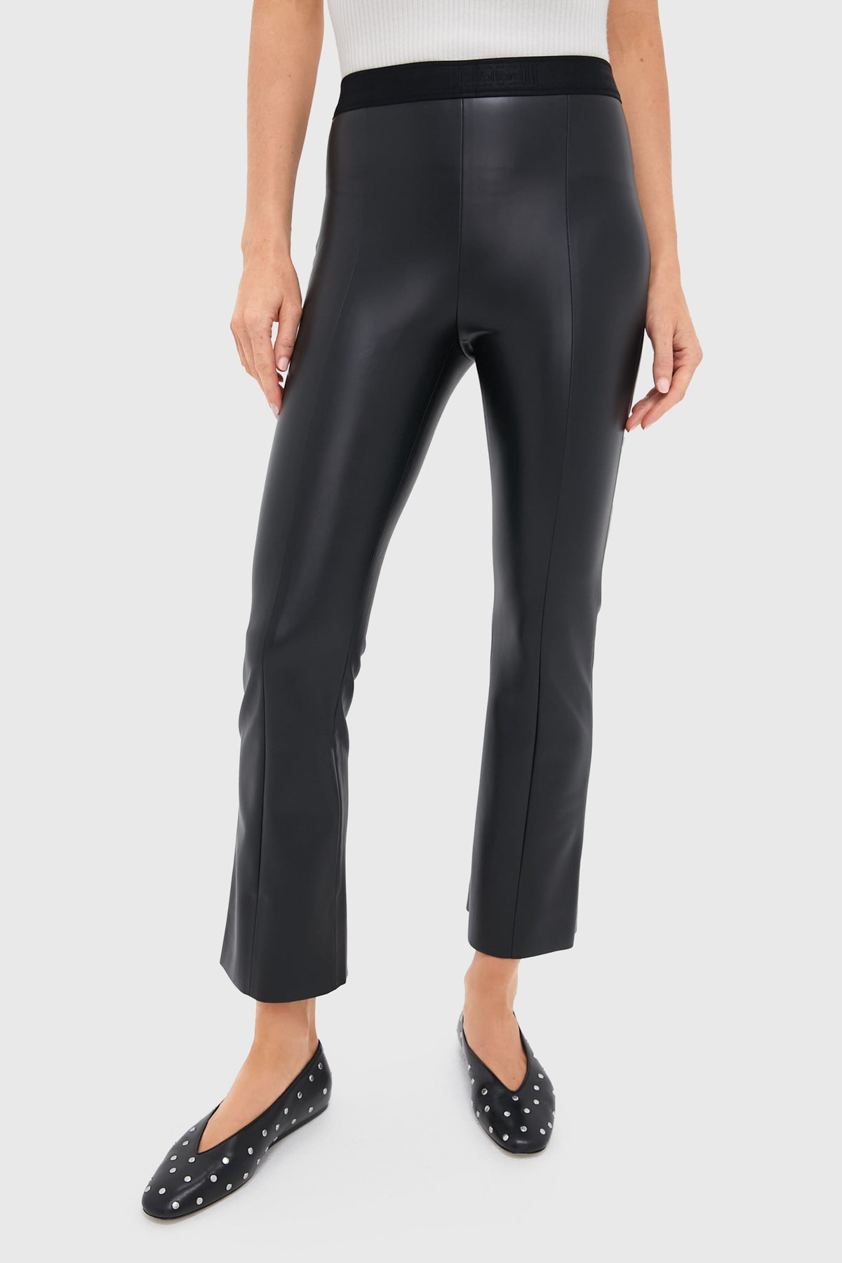 A person stands in a minimalist setting wearing black vegan leather trousers by Wolford and black studded flats.