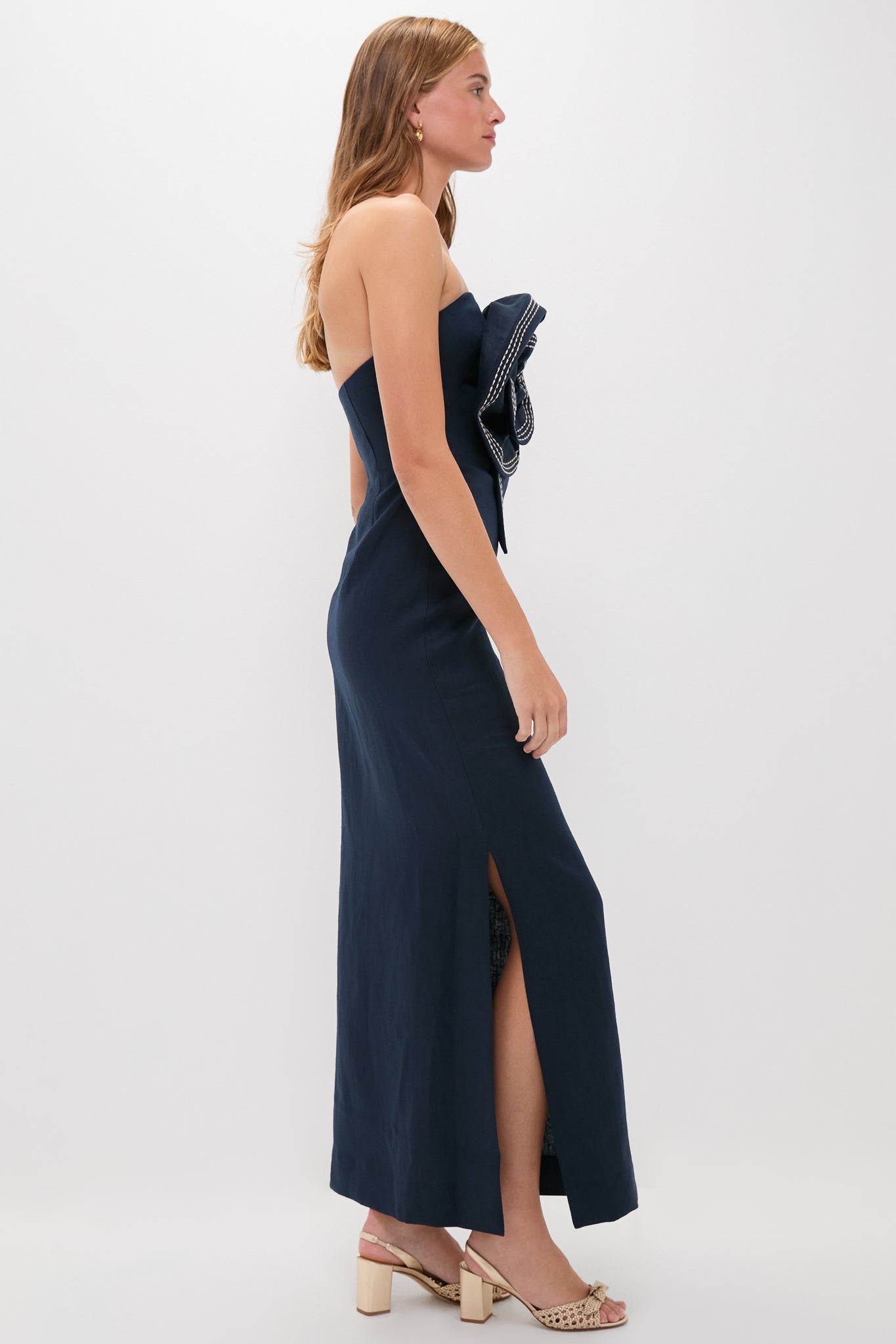 A woman in a navy blue, strapless midi dress with a side slit and large front bow detail, styled with beige heeled sandals.