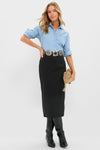 A woman models Hyacinth House: a light blue button-up shirt, high rise black column midi skirt, wide ornamental belt, black boots, and a tan clutch bag, all set against a plain white background.