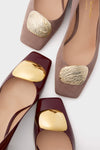 Close-up of two pairs of Rupert Sanderson women’s square-toe shoes: one taupe with textured gold-tone accents, the other in burgundy patent leather with smooth gold-tone embellishments.