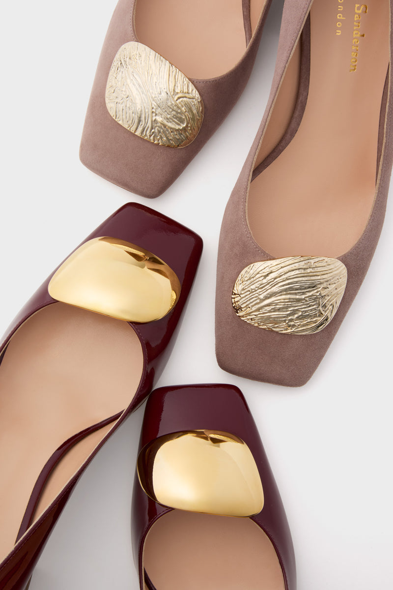 Close-up of two pairs of Rupert Sanderson women’s square-toe shoes: one taupe with textured gold-tone accents, the other in burgundy patent leather with smooth gold-tone embellishments.