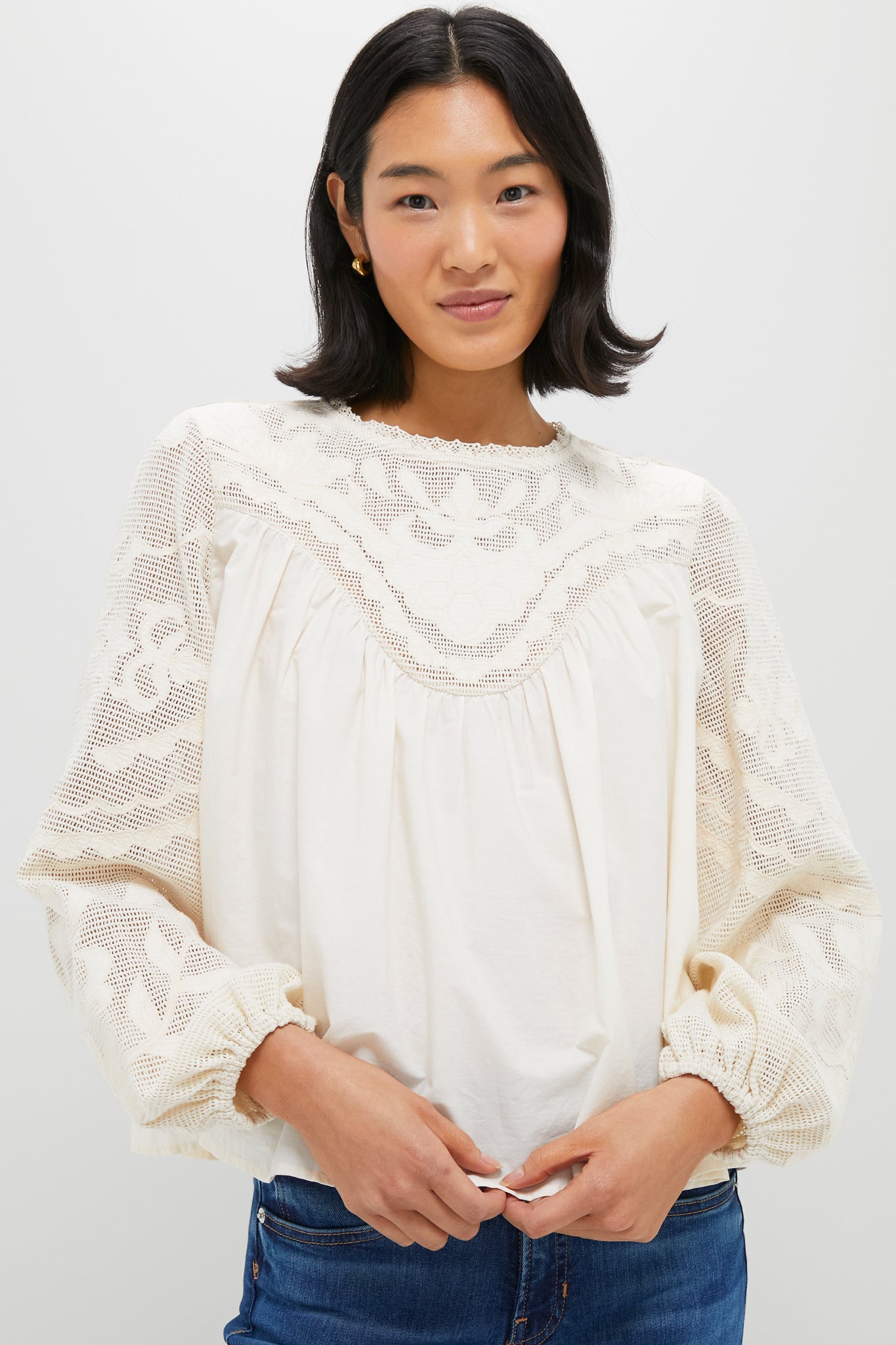 A person stands against a plain white background wearing a cream-colored Tuckernuck blouse with embroidered lace detailing on the sleeves and chest, and a relaxed fit.
