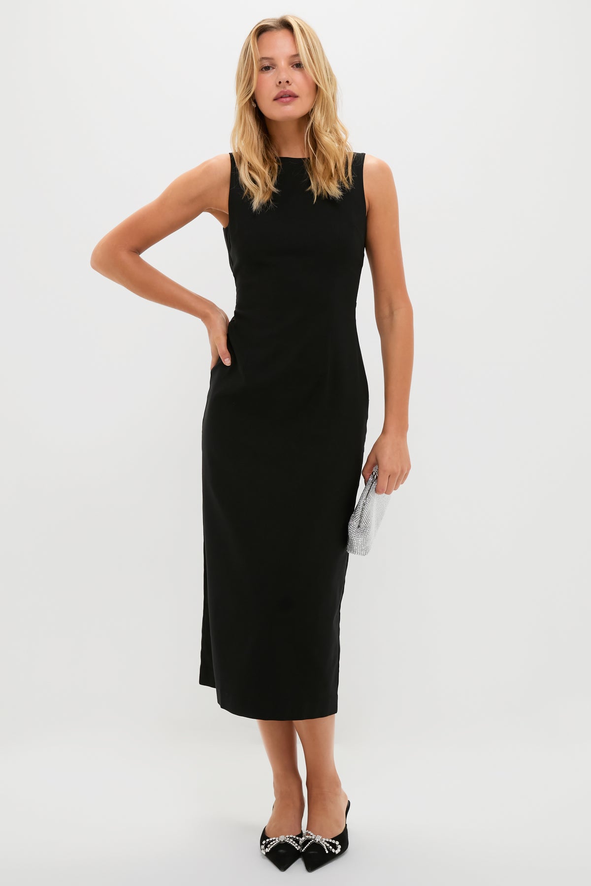 A woman poses against a plain white background wearing a Hyacinth House sleeveless black midi evening dress, black pointed bow heels, and holding a small silver clutch.