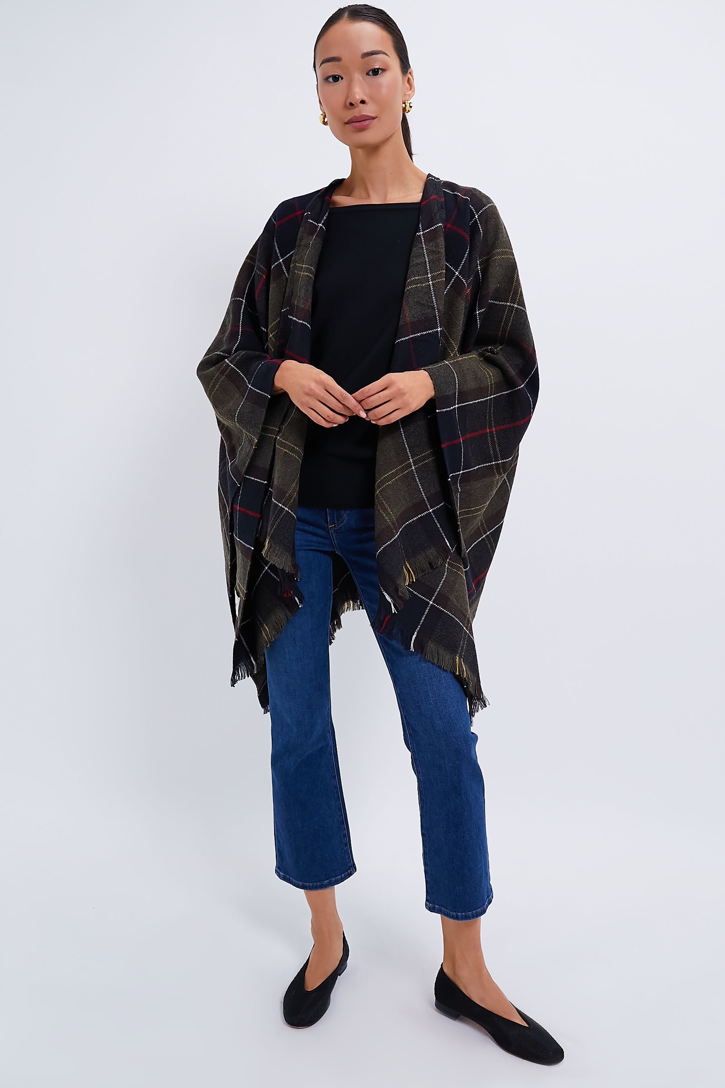 A woman models a Barbour plaid shawl over a black top, blue cropped jeans, and black flats, elegantly draping the stylish accessory around her shoulders.