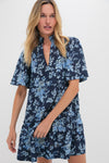 A woman wears the Tuckernuck Twilight Garden Crawford Dress, featuring a navy blue floral pattern, flutter sleeves, and a flattering tiered fit.