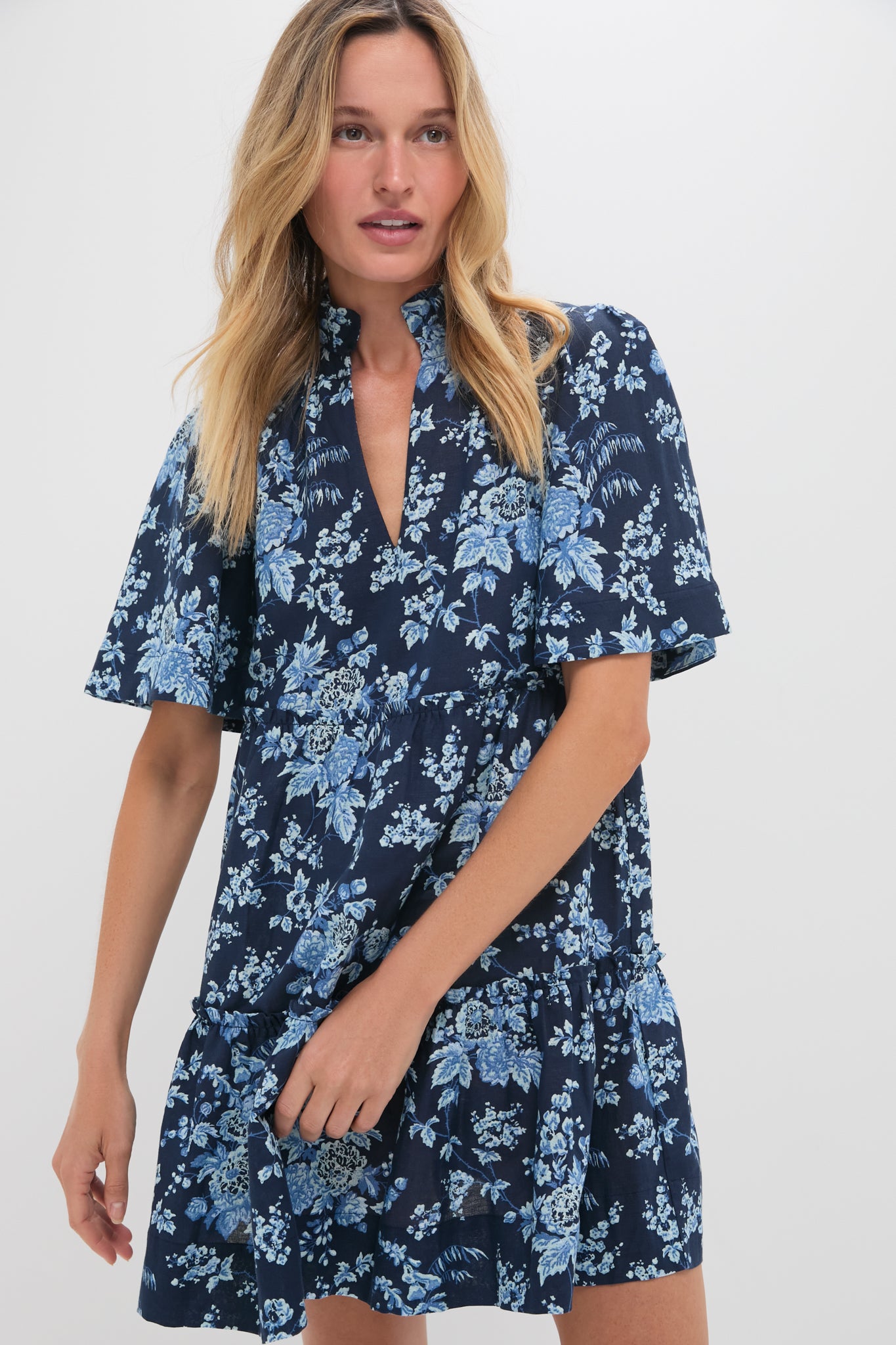 A woman wears the Tuckernuck Twilight Garden Crawford Dress, featuring a navy blue floral pattern, flutter sleeves, and a flattering tiered fit.