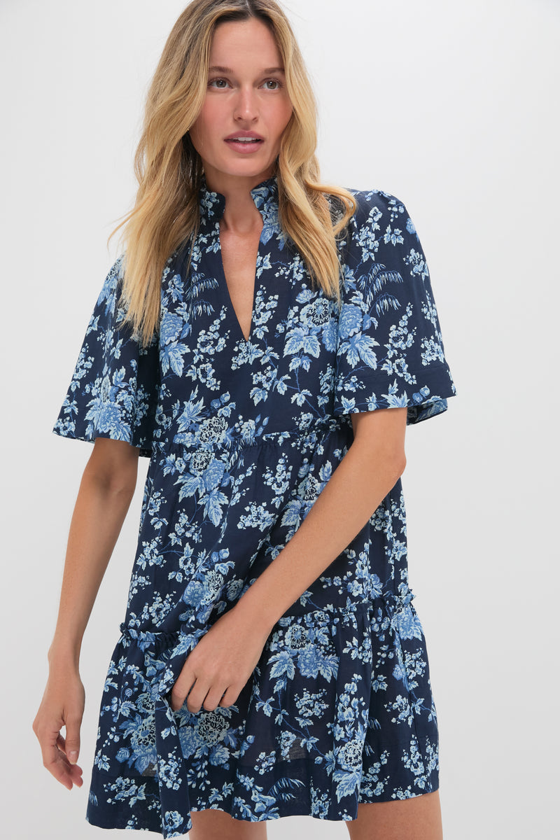 A woman wears the Tuckernuck Twilight Garden Crawford Dress, featuring a navy blue floral pattern, flutter sleeves, and a flattering tiered fit.