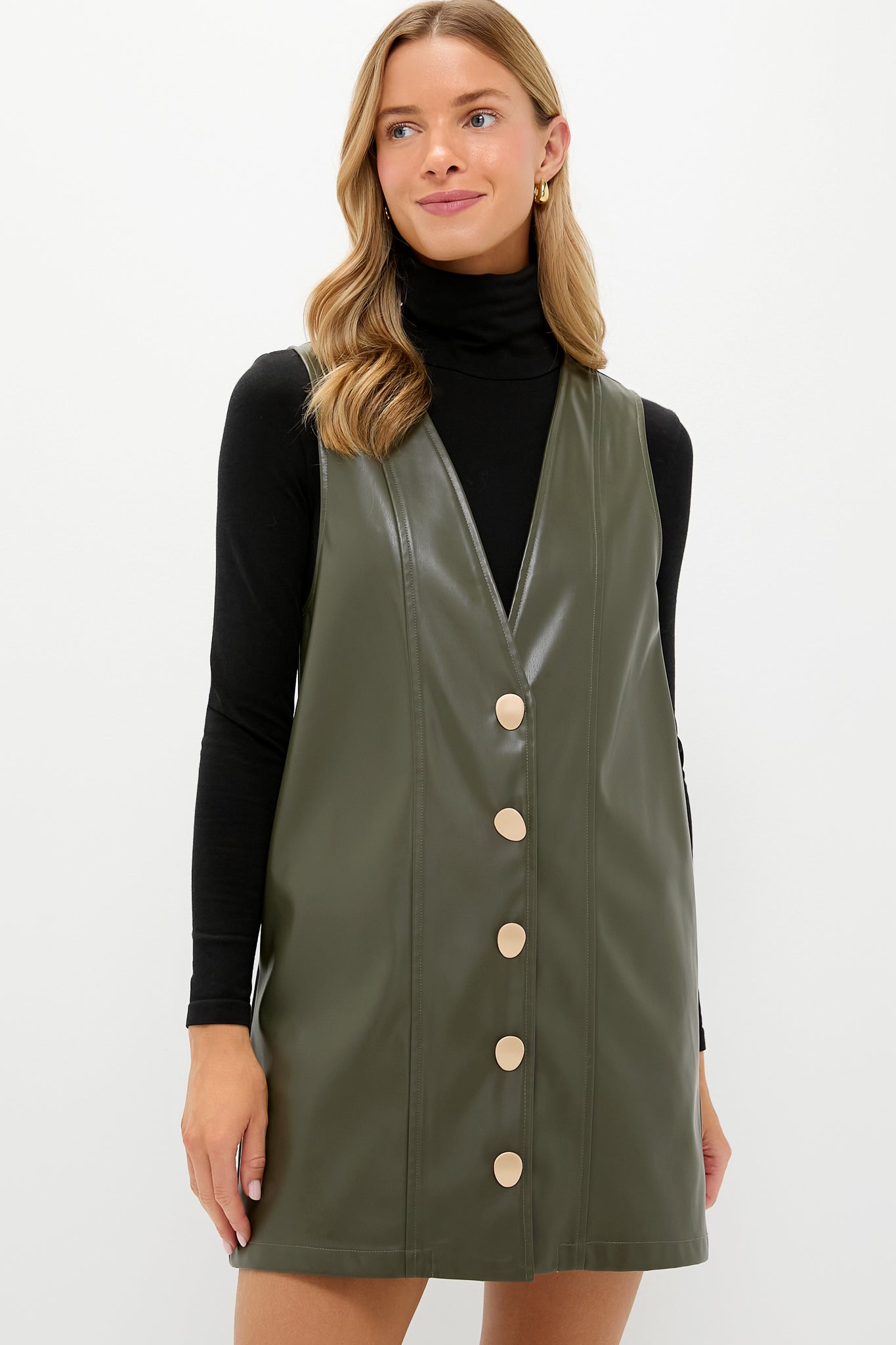 A woman models Hyacinth House’s olive green faux leather pinafore dress with large buttons, styled over a black turtleneck top.